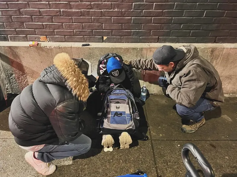 A Love on Hamilton volunteer kneeling on the sidewalk to care for a man sitting against a brick wall